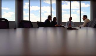 Business employees meet in a conference room with large windows revealing a partly cloudy afternoon.