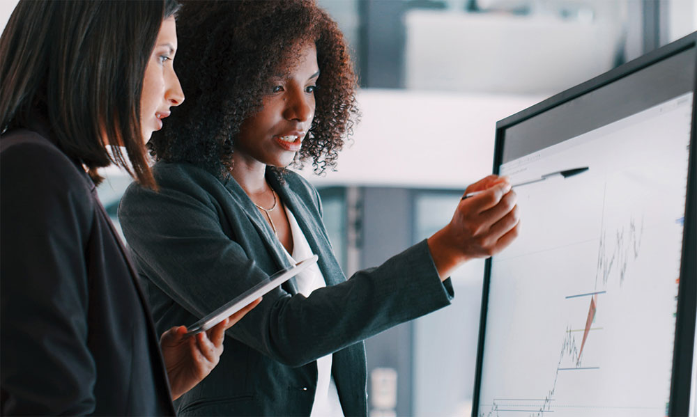 Two business analytics professionals have a discussion while looking at data graphs on a computer monitor.