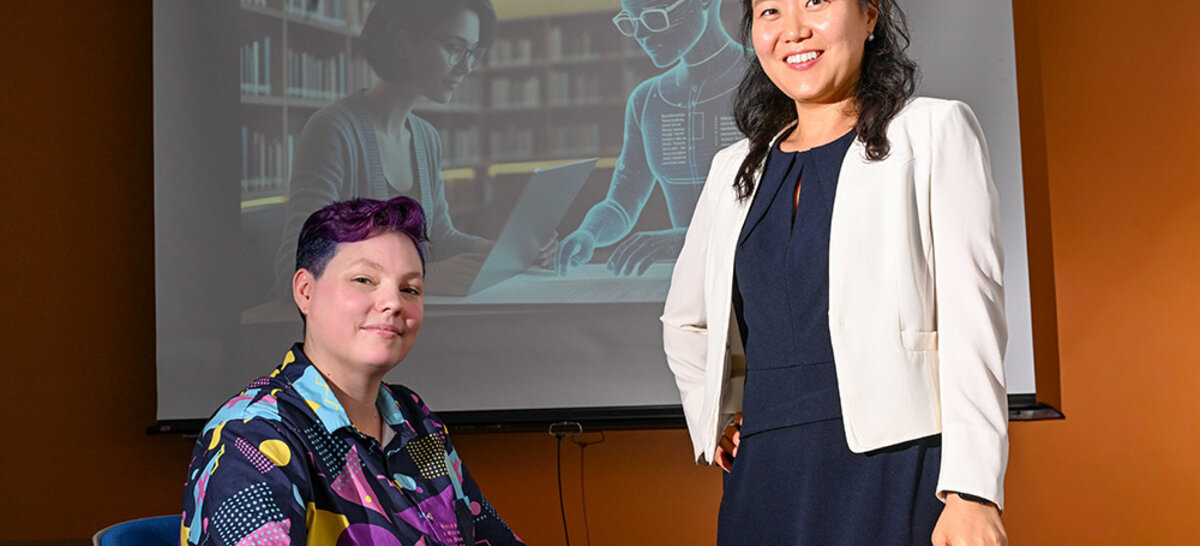 Dr. Jinhee Kim and PhD candidate Rita Detrick pose for a portrait in front of a screen with a presentation about AI.