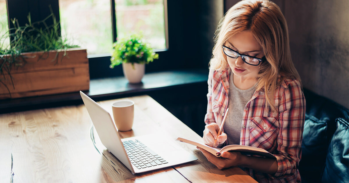 A woman studies in an online degree program while seated at a table with an open laptop and a notebook.