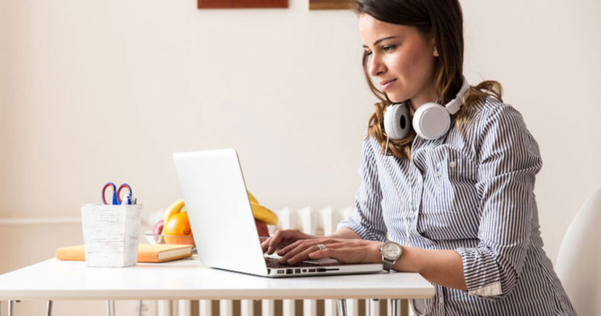 Woman with headphones around her neck types on a laptop seated at a kitchen table.