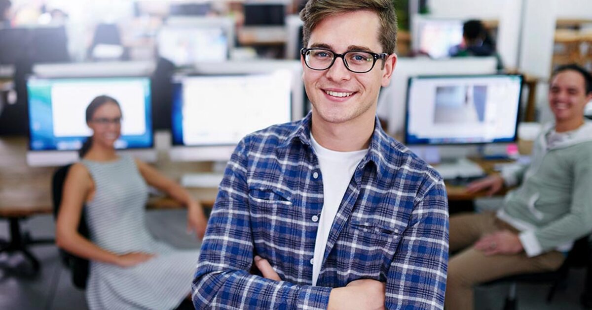 Young college student stands in an airy computer lab. His arms are folded and he is smiling at the camera.