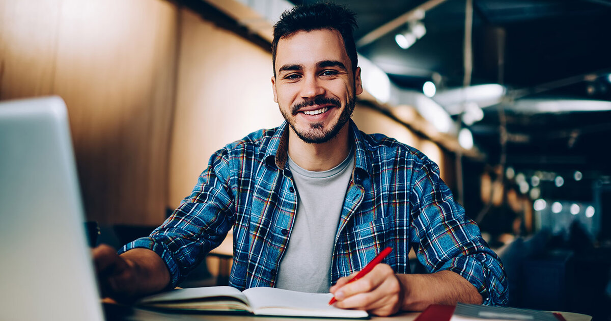 Smiling student sits at computer desk with a laptop and paper notebook