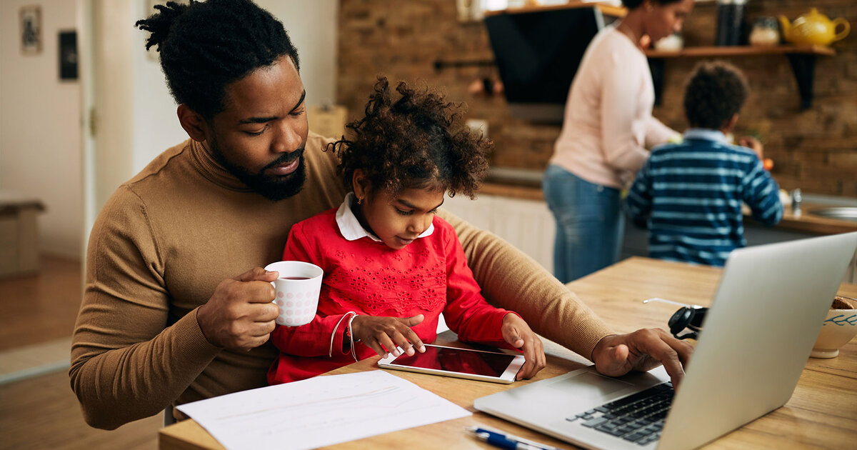 Dad works to transform his future with an online PhD program from ODUGlobal. While he sits working on his laptop his child looks at the screen and his family is in the background.
