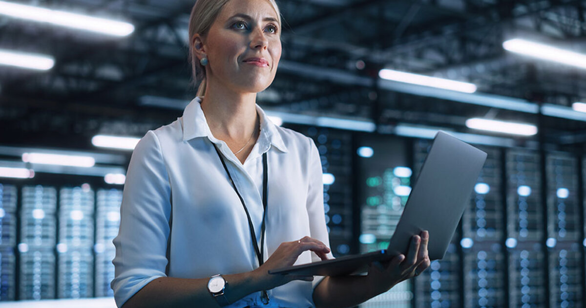 A data science professional stands holding a laptop, with server racks in the background