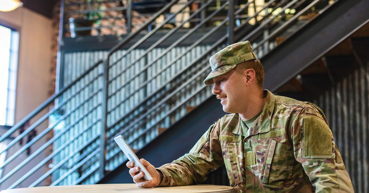 A seated active duty military service man looks at a handheld tablet device.