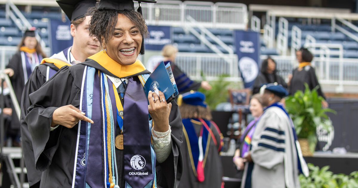 Athena Jones at Commencement with her diploma, excited and smiling at the camera while pointing to her oduglobal stole