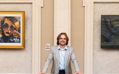 Current ODUGlobal student Caleb Lane stand in a local library surrounded by art and smiling at the camera.