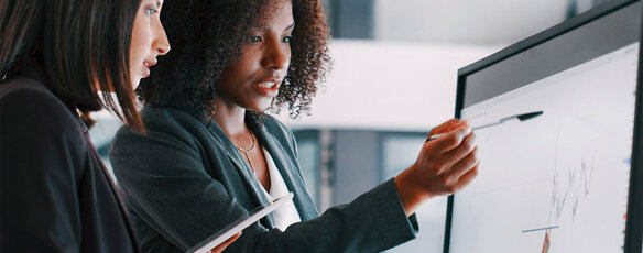 Two business analytics professionals have a discussion while looking at data graphs on a computer monitor.