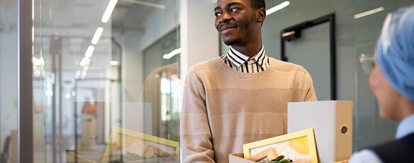 A smiling, young African American man arrives at his new job holding a box of items for his new office.