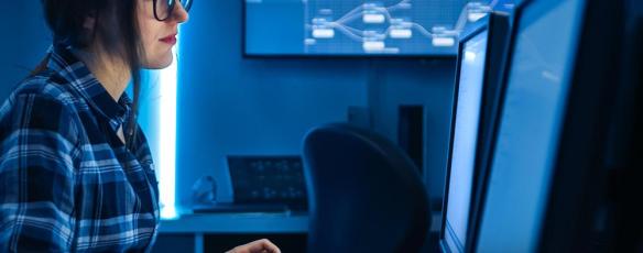 A female professional in the cybersecurity field works at a computer on a desk with dual monitors.