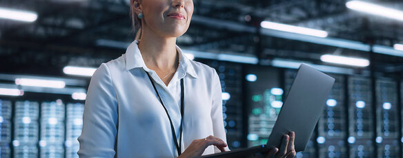 A data science professional stands holding a laptop, with server racks in the background