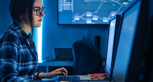 A female professional in the cybersecurity field works at a computer on a desk with dual monitors.