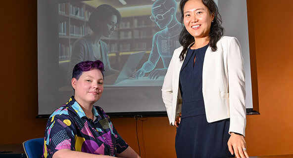 Dr. Jinhee Kim and PhD candidate Rita Detrick pose for a portrait in front of a screen with a presentation about AI.