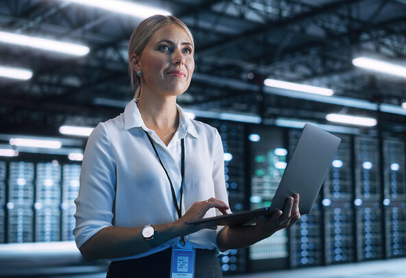 A data science professional stands holding a laptop, with server racks in the background