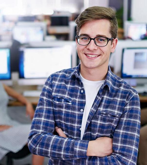 Young college student stands in an airy computer lab. His arms are folded and he is smiling at the camera.