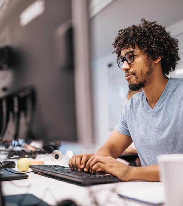 A young African American student is seated at a table in a computer lab, typing on a computer keyboard.