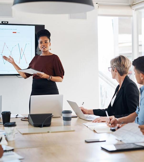 A data analytics professional stands by a chart and presents information to business leaders in a bright conference room.