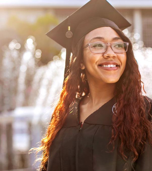 An ODU graduate wearing a graduation cap and gown smiles in the sunlight near ODU's lion fountain.