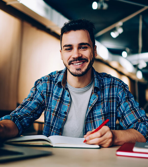 Smiling student sits at computer desk with a laptop and paper notebook