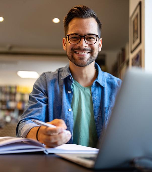 Student at desk with computer and books, smiling at the camera.