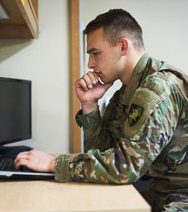 An active duty servicemember prepares for class at a laptop on a desk.