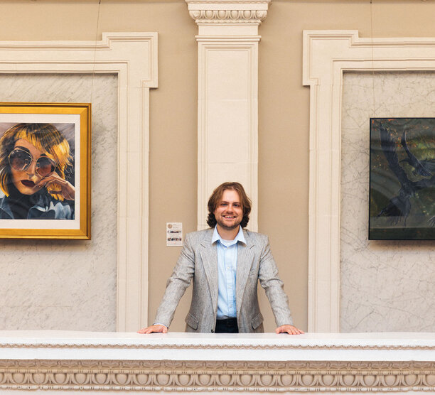 Current ODUGlobal student Caleb Lane stand in a local library surrounded by art and smiling at the camera.