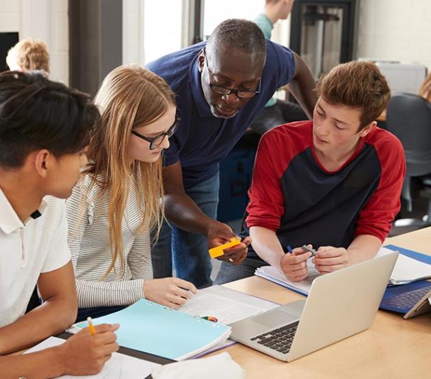 Technology education teacher leans over to talk to students seated at a table with a laptop and tablet