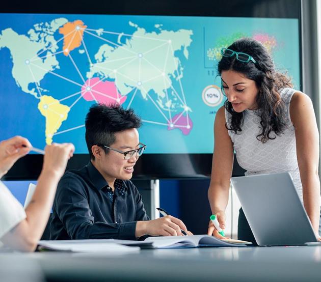 Geography professionals look at paperwork on a table with a world map on the large screen behind them.