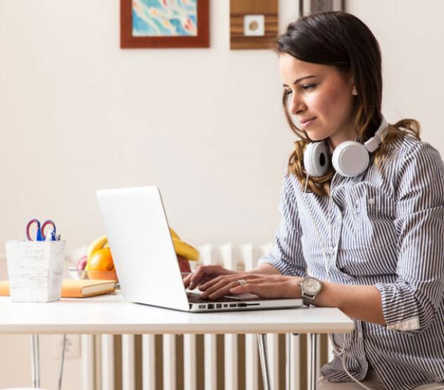 Woman with headphones around her neck types on a laptop seated at a kitchen table.