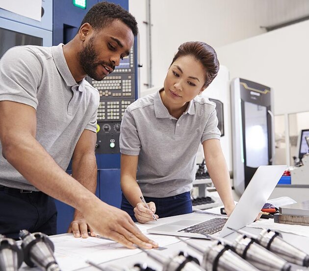 An industrial trainer and trainee review new processes at a laptop on a worktable with machine parts in the foreground.