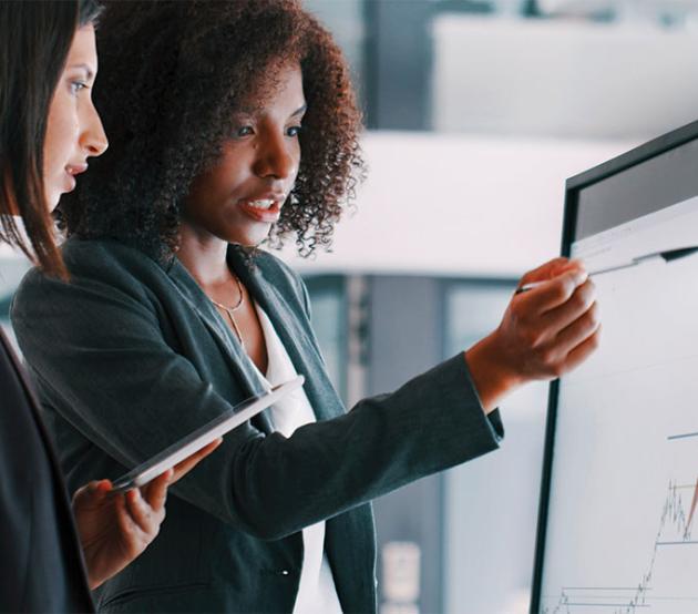 Two business analytics professionals have a discussion while looking at data graphs on a computer monitor.