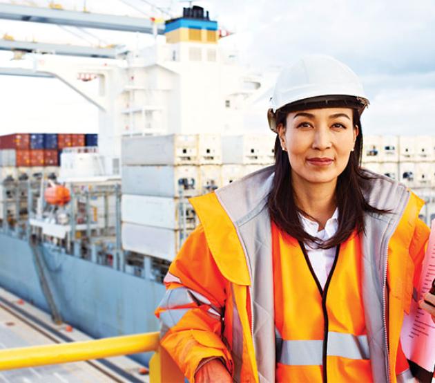 A woman in a hard hat and orange safety jacket stands on a balcony overlooking a cargo port where containers are being unloaded from a shipping vessel.