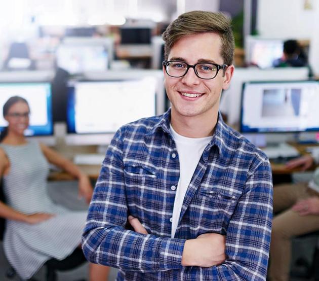 Young college student stands in an airy computer lab. His arms are folded and he is smiling at the camera.