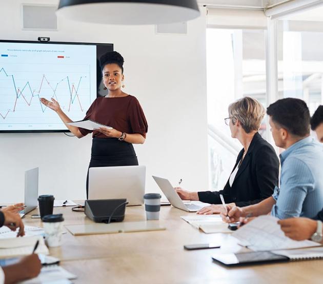 A data analytics professional stands by a chart and presents information to business leaders in a bright conference room.