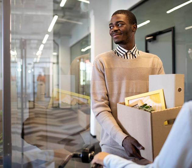 A smiling, young African American man arrives at his new job holding a box of items for his new office.