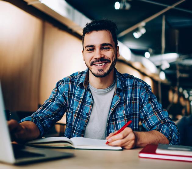 Smiling student sits at computer desk with a laptop and paper notebook