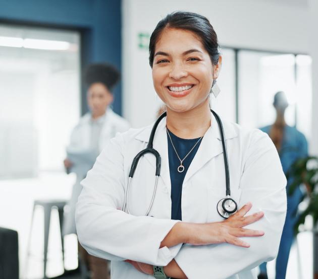 A smiling woman stands in a medical facility with arms crossed and a stethoscope around her neck