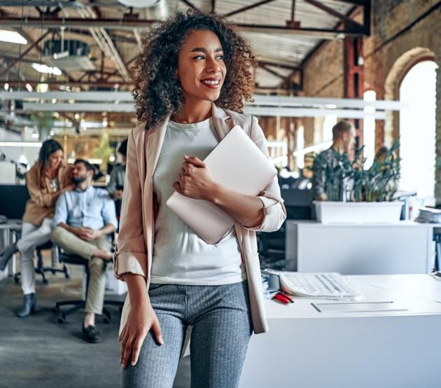 Woman stands in a professional workspace holding a laptop and smiling. She is headed to work on her online masters degree with ODUGlobal.