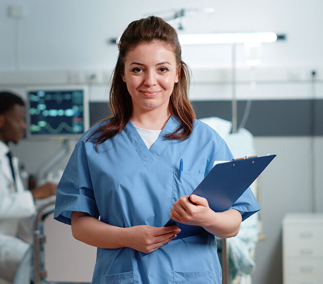 A nurse holding a medical chart stands in a patient's room and smiles with confidence.