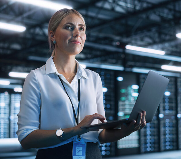 A data science professional stands holding a laptop, with server racks in the background