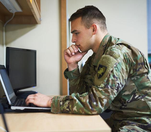 An active duty servicemember prepares for class at a laptop on a desk.