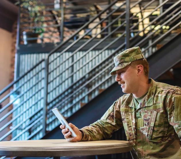 A seated active duty military service man looks at a handheld tablet device.