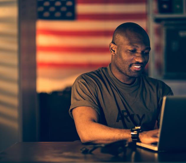 Military service member sits at a computer in front of the American flag