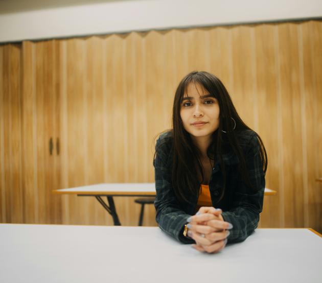 Woman sits at a desk in a classroom and looks at the camera intently 