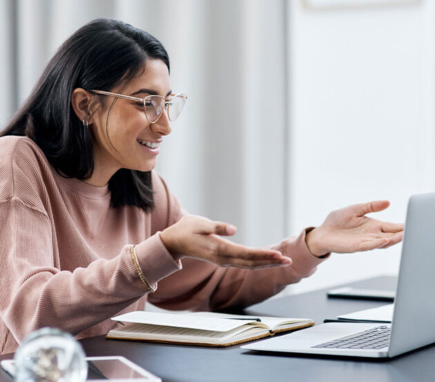 A young woman is seated at a table and conversing with others online through her laptop.