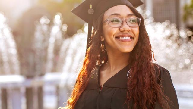 An ODU graduate wearing a graduation cap and gown smiles in the sunlight near ODU's lion fountain.