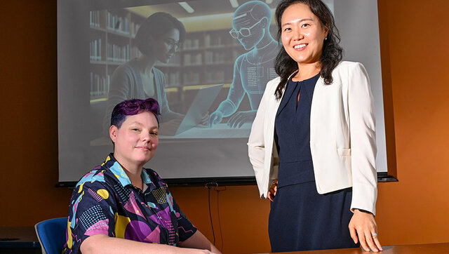 Dr. Jinhee Kim and PhD candidate Rita Detrick pose for a portrait in front of a screen with a presentation about AI.