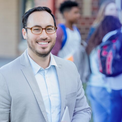 Man wearing a button shirt and sport coat smiles at the camera in a school hallway.