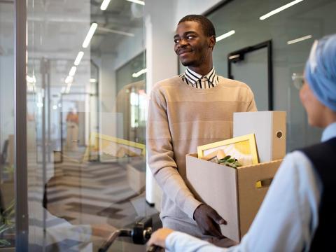 A smiling, young African American man arrives at his new job holding a box of items for his new office.